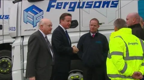 An archive picture showing former Prime Minister David Cameron, who is wearing a blue tie and a suit, shaking hands with a team of several workers from Fergusons Transport.