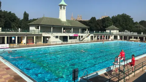 Orla Moore/BBC Peterborough Lido on a summer's day with a pool and pavilion buildings. In the foreground are two lifeguards in red on a raised platform.