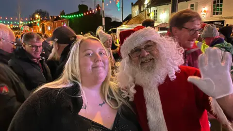 John Devine/BBC Devon Mandley, 32, has long blonde hair which is parted. She is wearing a black sparkly dress with a black cardigan on top. Some tattoos are visible near her neck. She is standing next to someone dressed as Santa, who is waving. Behind is a large crowd of people in the street with colourful lights strung over the road.