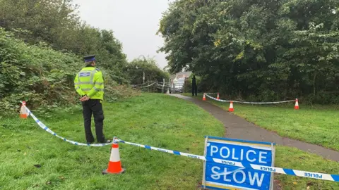 A police officer standing behind a line of police tape in a park
