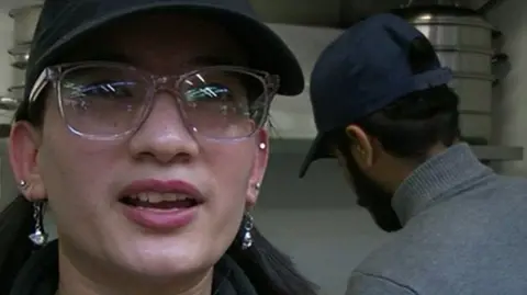 Close-up shot of Reeya Shresha. She is wearing glasses, earrings and a dark-coloured peaked cap. There is another person in the background preparing food.