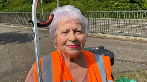 Margaret Faulkner has short grey hair with a pink tint. She has red lipstick and small gold earrings. She is wearing a high-vis orange tabard over a light pink V-neck top. She is carrying a litter grabber and the top of a rubbish bin is just behind her. Also in the background are some metal railings beside a roadway.