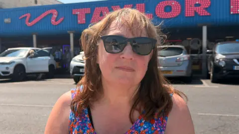 A lady in the sunshine with ginger hair, she's in a floral vest and is wearing sunglasses. She's at the sea front in Cleethorpes and she has a stern facial expression. 