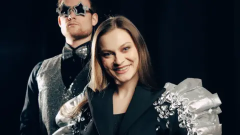 A dark haired girl, smiling at the camera, she is wearing a black blazer with silver ruffle detail and jewels on one shoulder