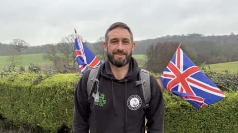 A man with dark hair and a dark beard wearing a black hoodie wears a grey rucksack on his back with two union jack flags coming from his back. He is stood in the countryside with a large hedge behind him.