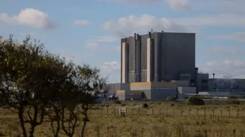 EDF Hartlepool nuclear power station is a tall, dark grey building surrounded by towers and pylons. There is smoke rising from some of the chimneys on the smaller, grey outer buildings. The facility is situated on a stretch of grass with a fence and foliage in the foreground, including the leafy top of a tree which is slightly out of focus.
