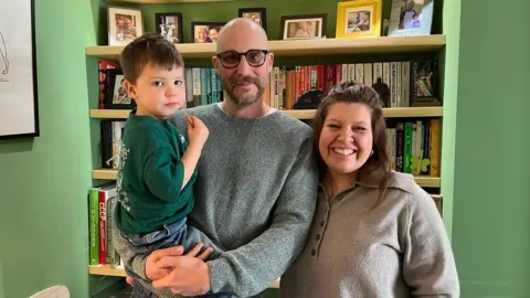 A family of three standing in front of a bookcase within a green wall. The man is in a grey jumper and black, round glasses. He is holding their son, who is holding a biscuit and wearing a green T-shirt. The woman is in a grey top, with shoulder length brown hair.