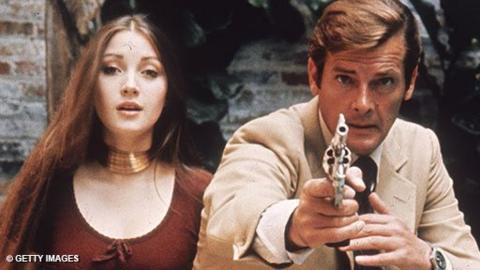 Jane Seymour as Solitaire in a red dress and gold necklace with surprised expression, alongside Roger Moore as James Bond in a light suit with magnum gun pulled.  {From 'Live And Let Die'}