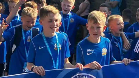 Glenavon FC youth players in a blue kits parade in Derry - there are two boys at the front holding a blue banner with the Glenavon badge on it. They are both wearing Nike-branded shirts with the Glenavon logo on them.