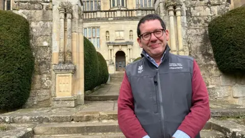 BBC/Seb Cheer Justin Scully smiles at the camera. He is wearing a grey gilet with the National Trust logo on it. He is standing in front of the grand facade of Fountains Hall.