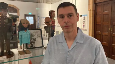 A man in a light blue shirt standing beside a display case of Thunderbirds puppets and memorabilia in a museum exhibition.