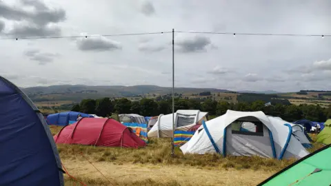 Tents pitched at the Kendal Calling campsite. The ground looks parched and the skies are grey. There is a tall pole propping up a string of lights across the site.