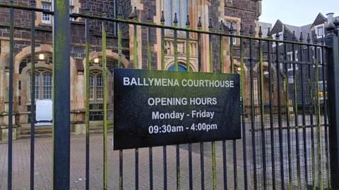 A black sign on black railings with white writing that says "BALLYMENA COURTHOUSE OPENING HOURS MONDAY - FRIDAY 09:30am - 4:00pm"