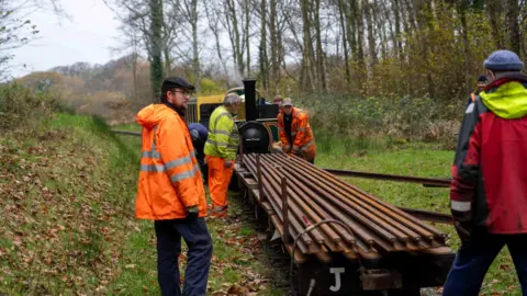 Ravenglass and Eskdale Railway Workmen load materials onto a flat trailer attached to a steam train. The train is pictured from behind with workmen around it. They are within woods along the track.