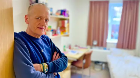 Man leaning against a door and looking at the camera. He is wearing a blue jumper and has several bracelets on his left wrist. He has short grey hair. In the background slightly blurred is a single bed on the left and on the right a chair and desk.