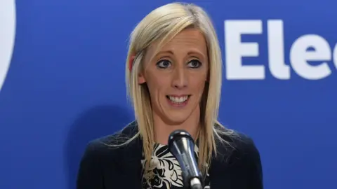 PA Media Carla Lockhart has straight, long blond hair and is wearing a black suit jacket and white and black scarf. She is speaking at an election event last yeat 