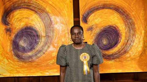 PA Media Nnena Kalu em frente a dois grandes desenhos com fundo laranja e formas espiraladas