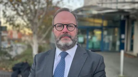 David Dadds, wearing black-framed glasses, a white shirt with a blue patterned tie and a grey suit jacket, standing in front of the entrance to Exeter City Council. He has grey hair and a full grey beard and he has a serious expression on his face.