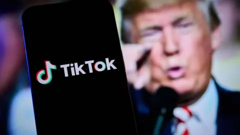 A phone showing the tiktok logo is seen in front of an image of Donald Trump pointing at a camera.