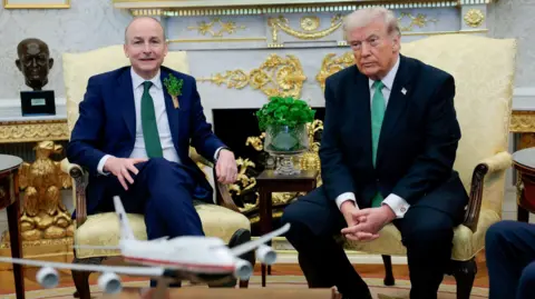Two men in navy suits, wearing green ties. They are sitting on yellow chairs in an ornate room. The man in the left has shamrocks in his suit pocket and has little hair. The man on the right has blonde hair and is wearing a US flag pin.