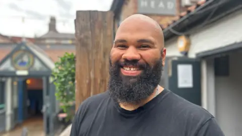 BBC/Jasmine Lowe Aiden Hazlewood, who has a bald head and black beards, smiles. Behind him is The King's Head pub