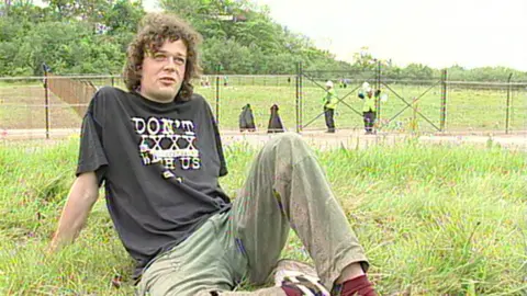 A man sat on gras leaning back onto his arms. He has curly brown hair and is wearing a black t-shirt and green trousers. Men in high visibility jackets are in the background next to a metal fence.