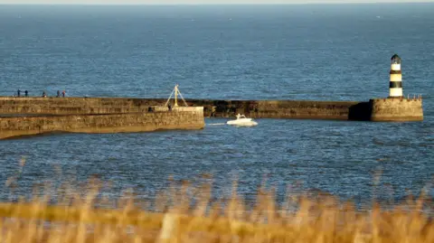BBC Weather Watchers/Dan Seaham Harbour Lad A small whit boat in between two stone piers. The one that curves further to the right has a small black and white striped lighthouse on the end. There are people walking along them. The sea is blue and calm and everything is cast in yellowish light. In the foreground of the photo there is some yellow grass.