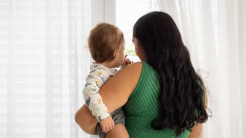 A woman in a green top and with long dark, wavy hair holding a toddler in her arms. They are looking out thw eindow through white curtains and facing away from the camera.