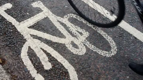 An image of a bicycle is drawn on the tarmac in white paint to denote a cycle path. On the right is the lower part of a bicycle wheel.