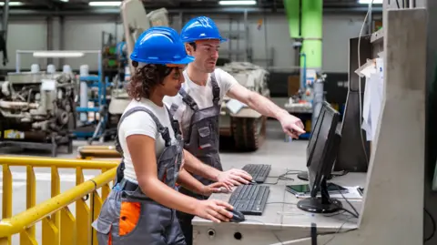 Two workers wearing blue hard hats and grey overalls operate computers at a metal workstation on a factory floor. One worker points at a monitor while the other uses a keyboard and mouse. Industrial machinery, a yellow safety railing, and production equipment are visible in the background.