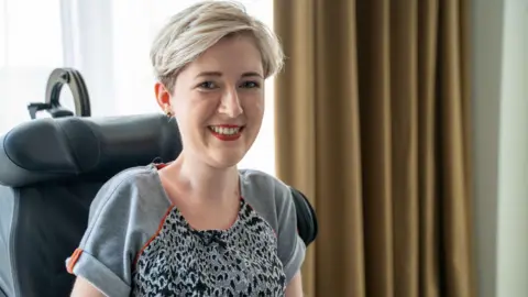 Sarah Rennie Sarah Rennie has short blonde hair and is sitting in her wheelchair, smiling at the camera. She is wearing a grey leopard print t-shirt and red lipstick