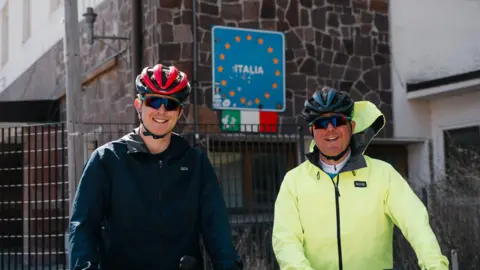 Family Handout Josh and George Kohler are standing side by side and in front of a building which has a blue sign that reads ITALIA. The are both wearing cycling helmets, sunglasses and coats.
