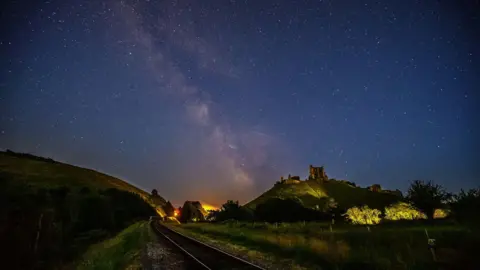 Richard Murgatroyd FRIDAY - The night sky over the Dorset countryside. A railway line runs through the centre, with the ruins of Corfe Castle visible on a hill to the right with a green hill to the right of the tracks It is night so the colours are muted, but in the distance you can see the glow from the street lights in Corfe Castle village. In the dark sky above there are thousands of stars with the milky way running through the centre of the sky.