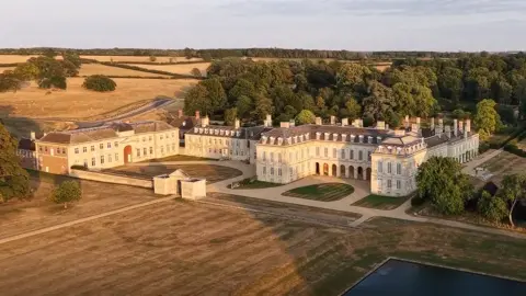 Boughton House The outside of a stately home, showing a large building, in several sections, with trees behind it, lawns in front of it, and a an area of water to the right. The building has a large slate roof. Fields are also to the left. 