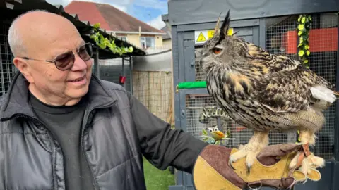 A man in a black jumper and puffer jacket smiles in a garden, as an owl sits on his hand in a bird handling glove