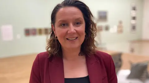 Stuart Whincup/BBC Dr Laura Sillars, Director of MIMA, smiles at the camera in one of the rooms. She's wearing a red suit and black top and the background is blurry. It looks like art or dance studio with a brown, wooden floor. 