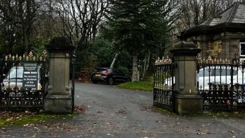 Stone pillar entrance gates to Crossley Heath School in Calderdale, with the top (finials) missing after having been stolen. The black and gold gates are open, with parked cars, grass and trees either side of the driveway.