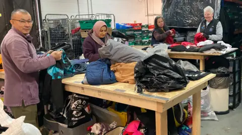 A man and three women are working to sort clothes set out of two wooden tables in a warehouse.