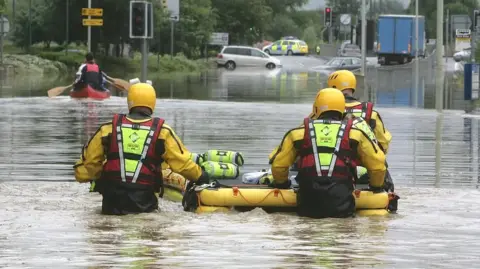 Getty Images Four men in yellow wetsuits using an inflatable raft in flood water