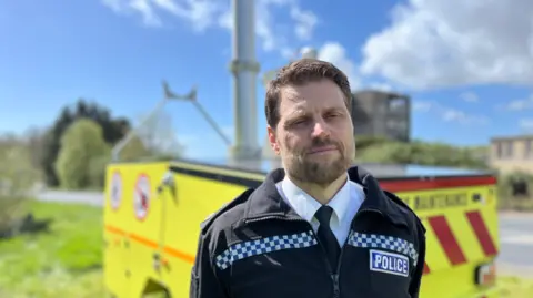 A police officer in uniform is standing in front of a bright yellow AI camera by the side of the road, with a blue sky and trees behind