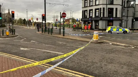 Kevin Fitzpatrick/BBC A yellow police tape runs across a road in Oldham with emergency services seen gathering in the distance. 
