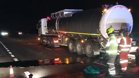 National Highways Tanker with black liquid spilling on to a motorway - two highways worked stand at the back of the tanker - it is night.