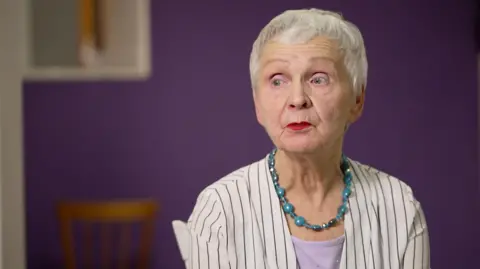 Ludmila Vasilyeva, 84, sits in a black and white vertical striped shirt, a turquoise bead necklace and pink shirt, facing camera left in front of a purple wall