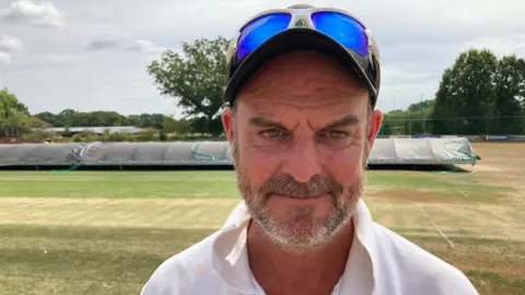 A man with a cap on, with blue tinted sunglasses on top of the cap. He has brown and grey facial hair and is wearing a white t-shirt. In the background is a cricket pitch with brown and green grass, and black rain covers