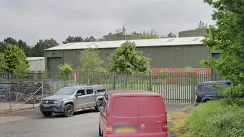 Google A green corrugated metal building stands behind green metal fences. The car park is empty. In front of the fence and gate are three vehicles