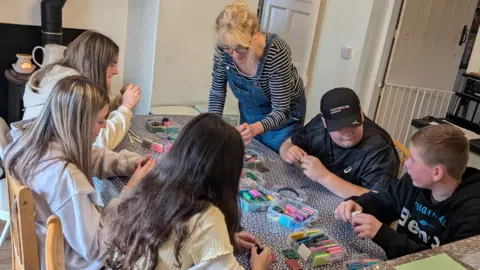 Nest Pembrokeshire Three teenage girls and two teenage boys sat around a kitchen table. The girls have long brown hair, one of the boys has short fair hair and the other is wearing a black baseball cap. Boxes of coloured model clay are in the middle of the table and they are all chatting and moulding the clay. A blonde woman wearing a striped t shirt and denim dungarees stand next to them and leans over the table. 