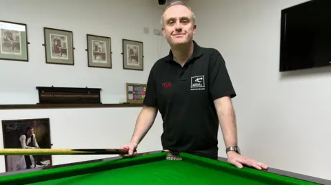 BBC Aaron Canavan, a man with grey hair, wearing a black polo shirt with red and white badges on the chest. He is holding a snooker cue and leaning over a green snooker table, while staring directly at the camera. Behind him are black and white, framed photos arrayed on the wall. There is black television on the wall over his left shoulder.