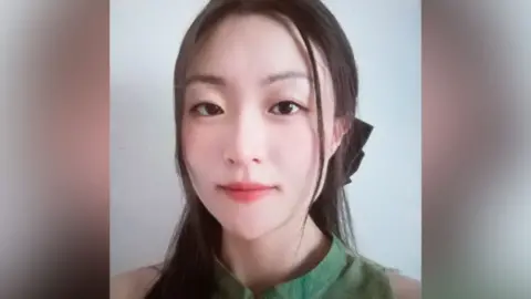 Met Police Picture of Zhe Wang wearing a green top. Her dark straight hair is tied back in a pony tail and she is looking straight at the camera.