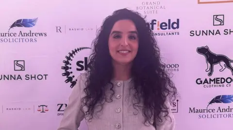 BBC A woman with curly hair stands in front of a board smiling into the camera.