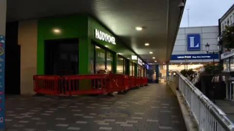 Bedfordshire Police An outdoor ramp leading up to Luton Point shopping centre. There are grey and white bricks on the floor and a metal railing on the right. There is a row of shops on the left of the ramp, the main one in shot is Paddy Power with a green exterior and white sign. It is quite gloomy with a grey sky.